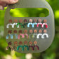 A hand holds a translucent olive-green display board showing multiple colorful U-shaped earrings with round copper-colored backing discs, against a blurred green outdoor background.