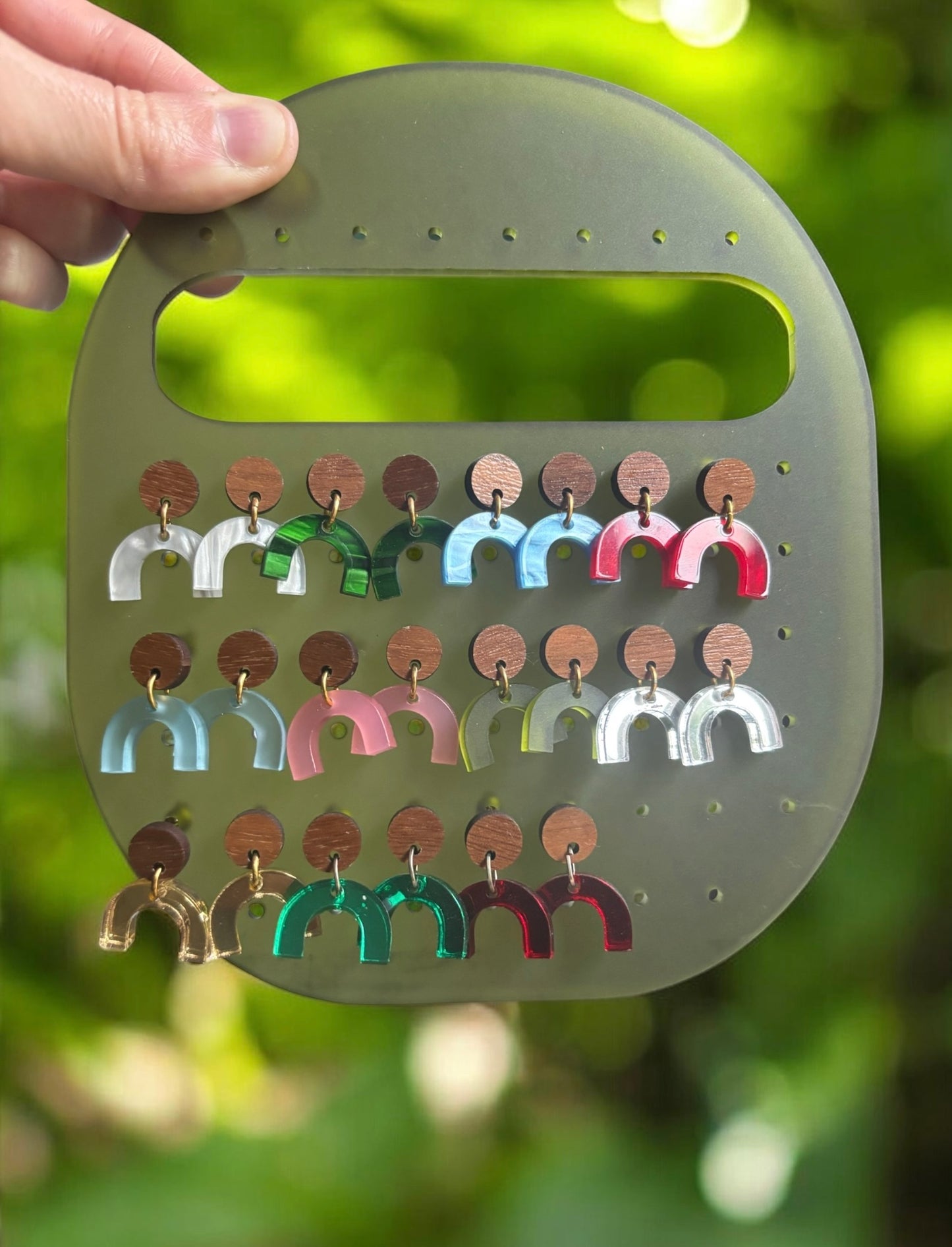 A hand holds a translucent olive-green display board showing multiple colorful U-shaped earrings with round copper-colored backing discs, against a blurred green outdoor background.