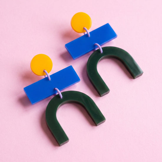 A pair of geometric drop earrings on a solid pink background. Each earring features a bright blue rectangular bar, a small yellow circular stud, and a dark green U-shaped drop.