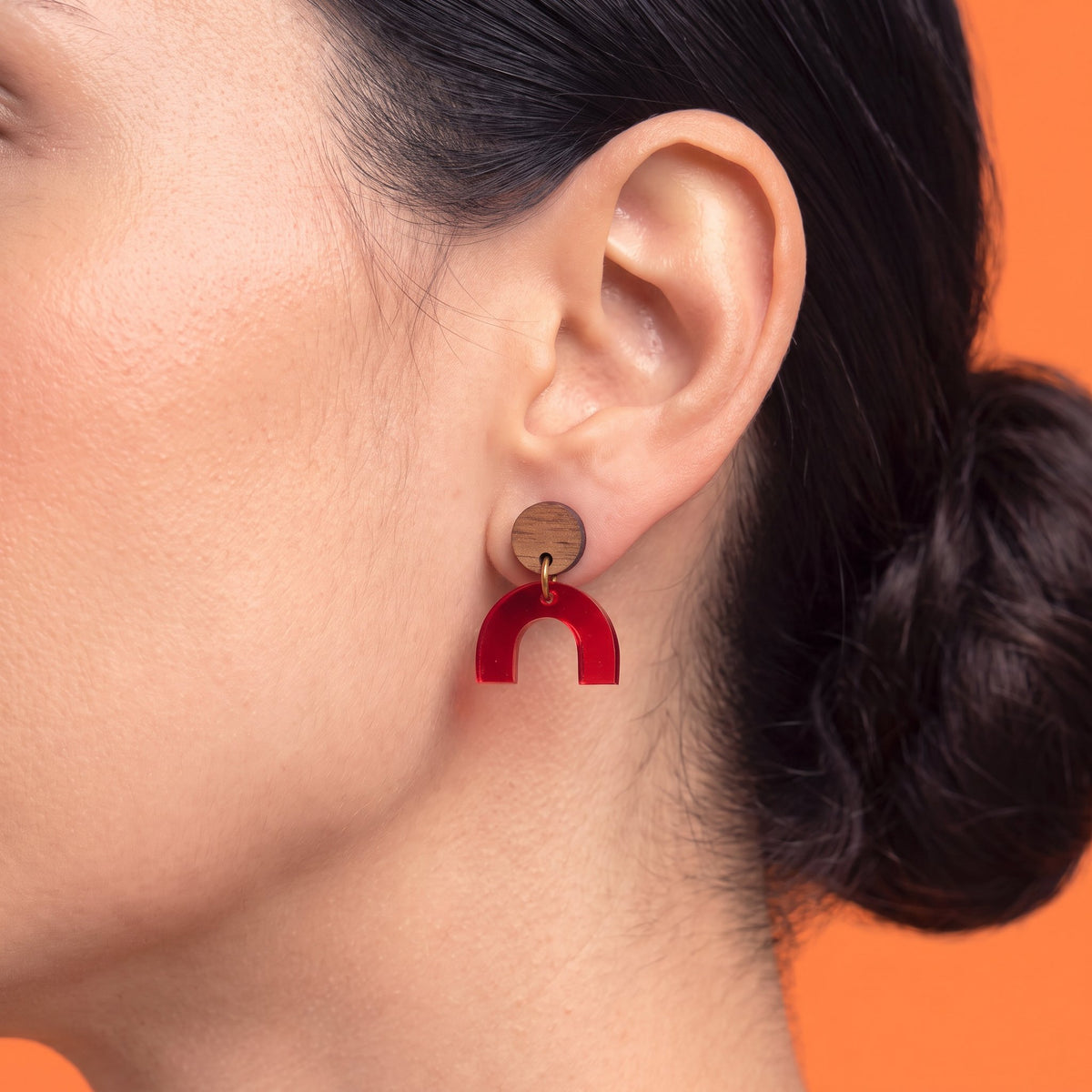 A close-up of a woman's ear wearing a wooden stud earring with a red U-shaped acrylic drop, set against a solid orange background.