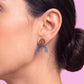 A close-up of a woman's ear wearing a wooden stud earring with a blue-gray arch-shaped drop, set against a solid pink background.