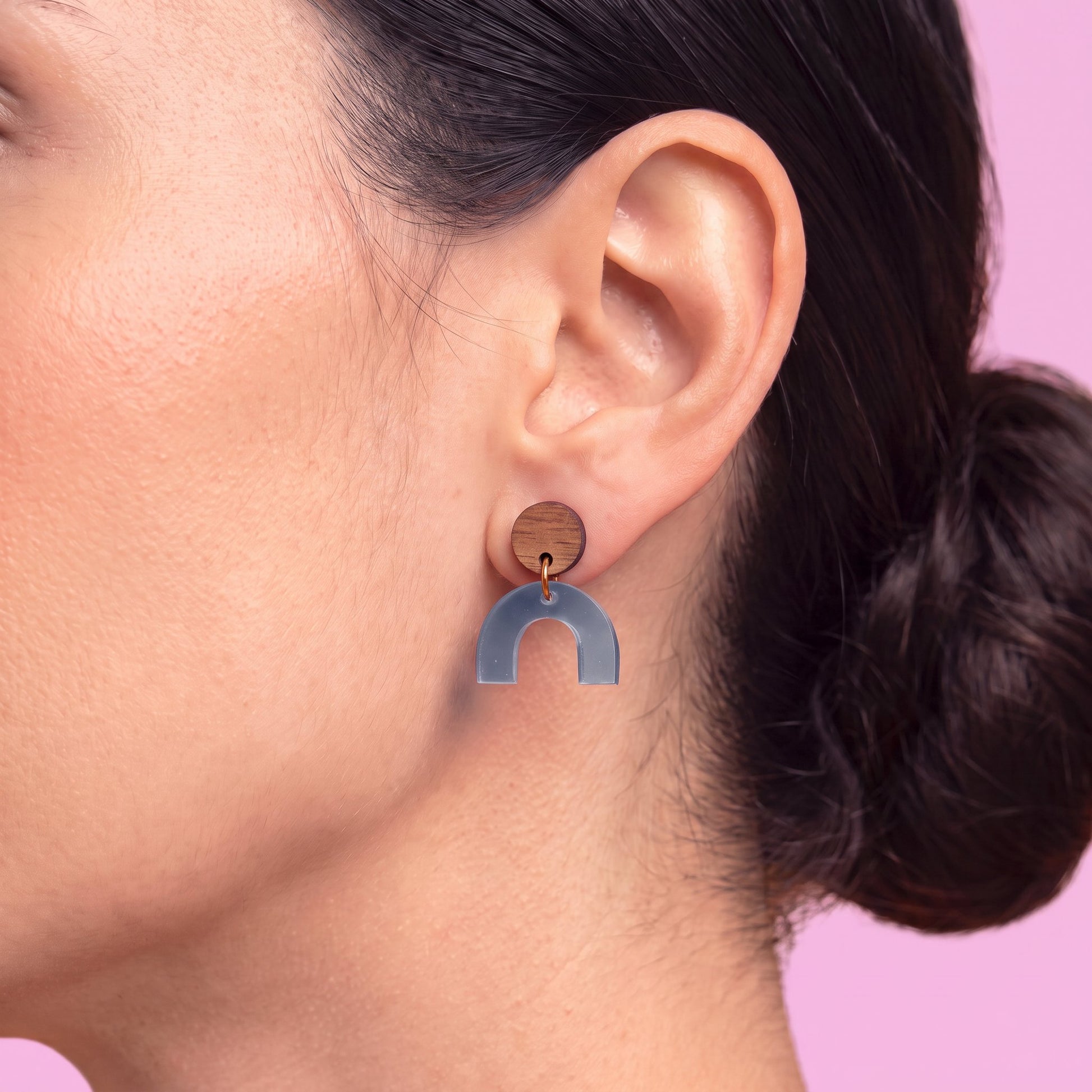 A close-up of a woman's ear wearing a wooden stud earring with a blue-gray arch-shaped drop, set against a solid pink background.