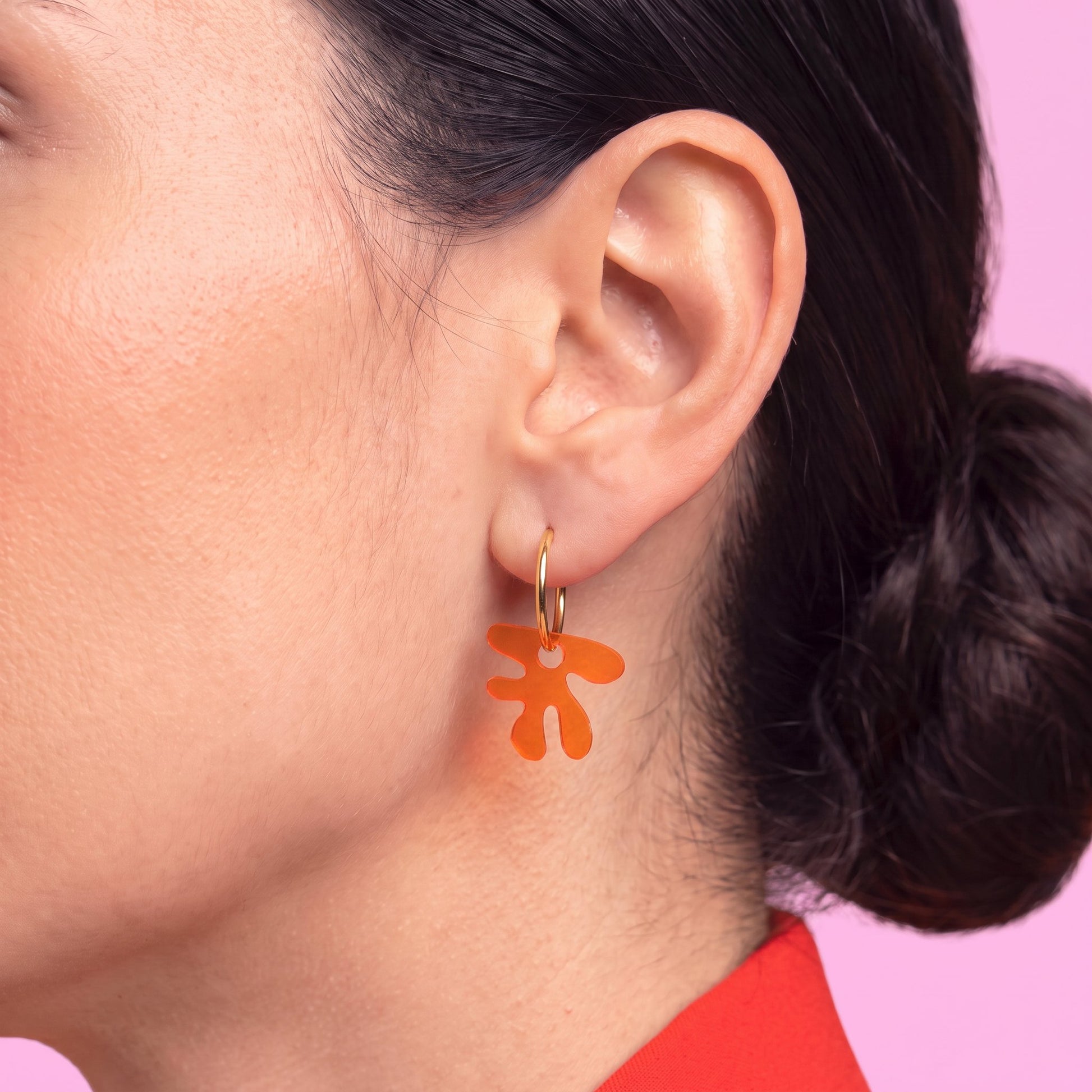 A close-up of a woman's ear wearing a gold hoop earring with a dangling orange flower-shaped charm, set against a solid pink background.